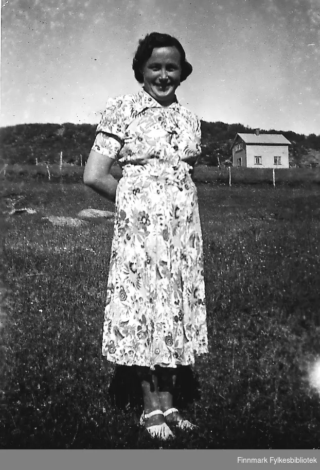 En kvinne, Amalie Mathisen, ståe på en eng. Dagen er solig og fin, hun er kledd i en lys blomstret sommerkjole.
