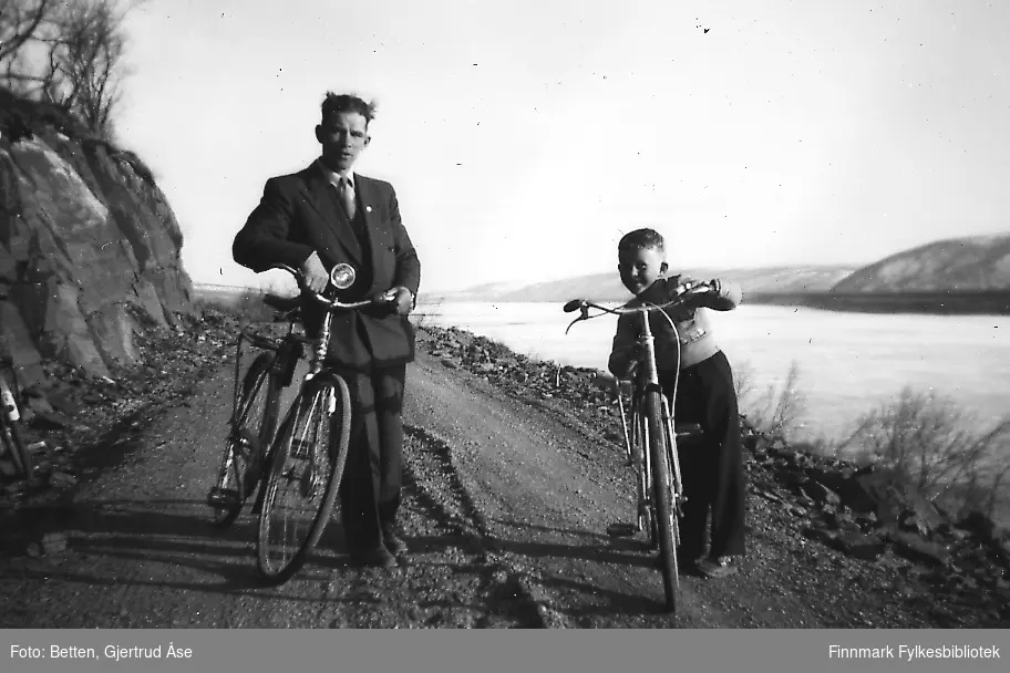En mann og en gutt på sykkeltur 17. mai i 1956. Vaktmester Anreassen fra Seida skole og internat og Bjørn Ottar Betten står med syklene sine ved Tanaelva som ennå er dekt av is. Andreassen har på seg en mørk dress og slips.