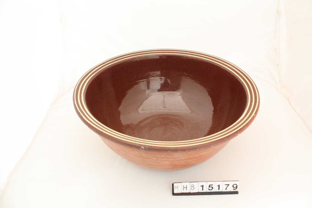 Brun keramikkskål, med glasert innside og ubehandlet utside. Munningskanten har dekor med tre parallelle gule streker som følger kanten rundt.