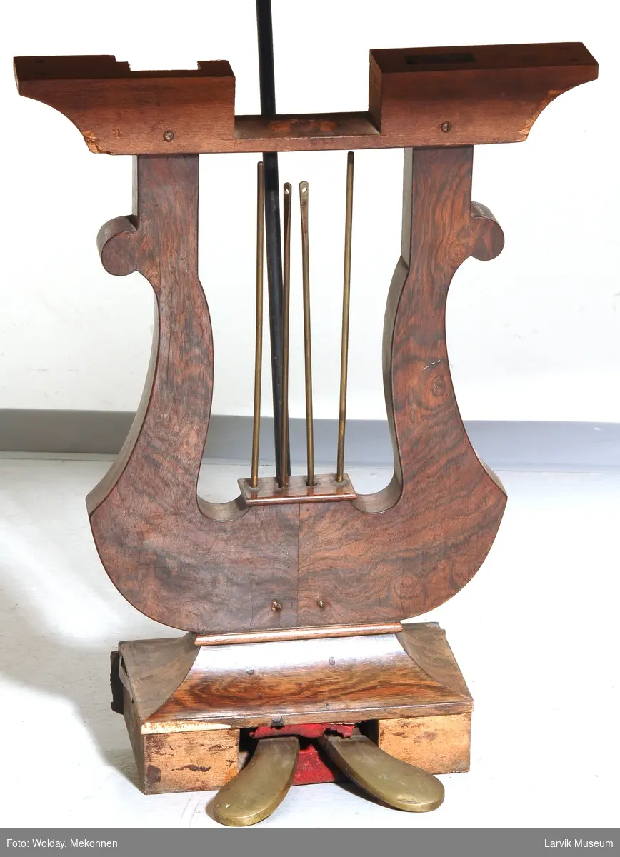 Piano i form av rektangulær kasse i mahogni med hengslet lokk. Profilerte ben med hjul under hvert ben. Lyreformet forbindelse mellom pedaler og kasse. 
