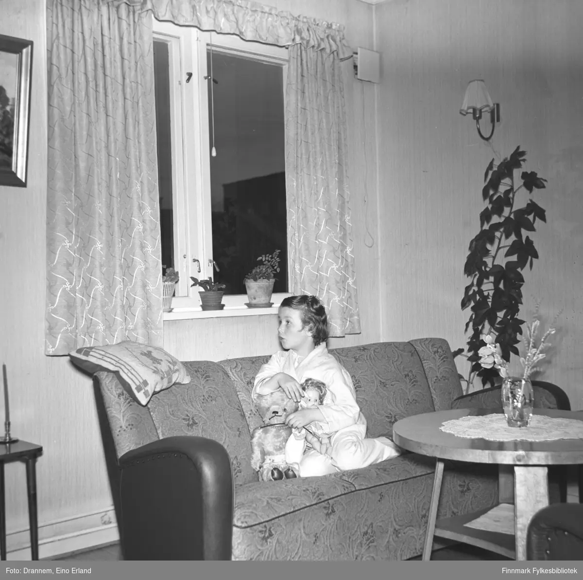 Turid Lillian sitter i sofaen, hun har en dokke og bamse på fanget sitt. Bildet er tatt i leiligheten til familien Drannem i Tollboden i Hammerfest