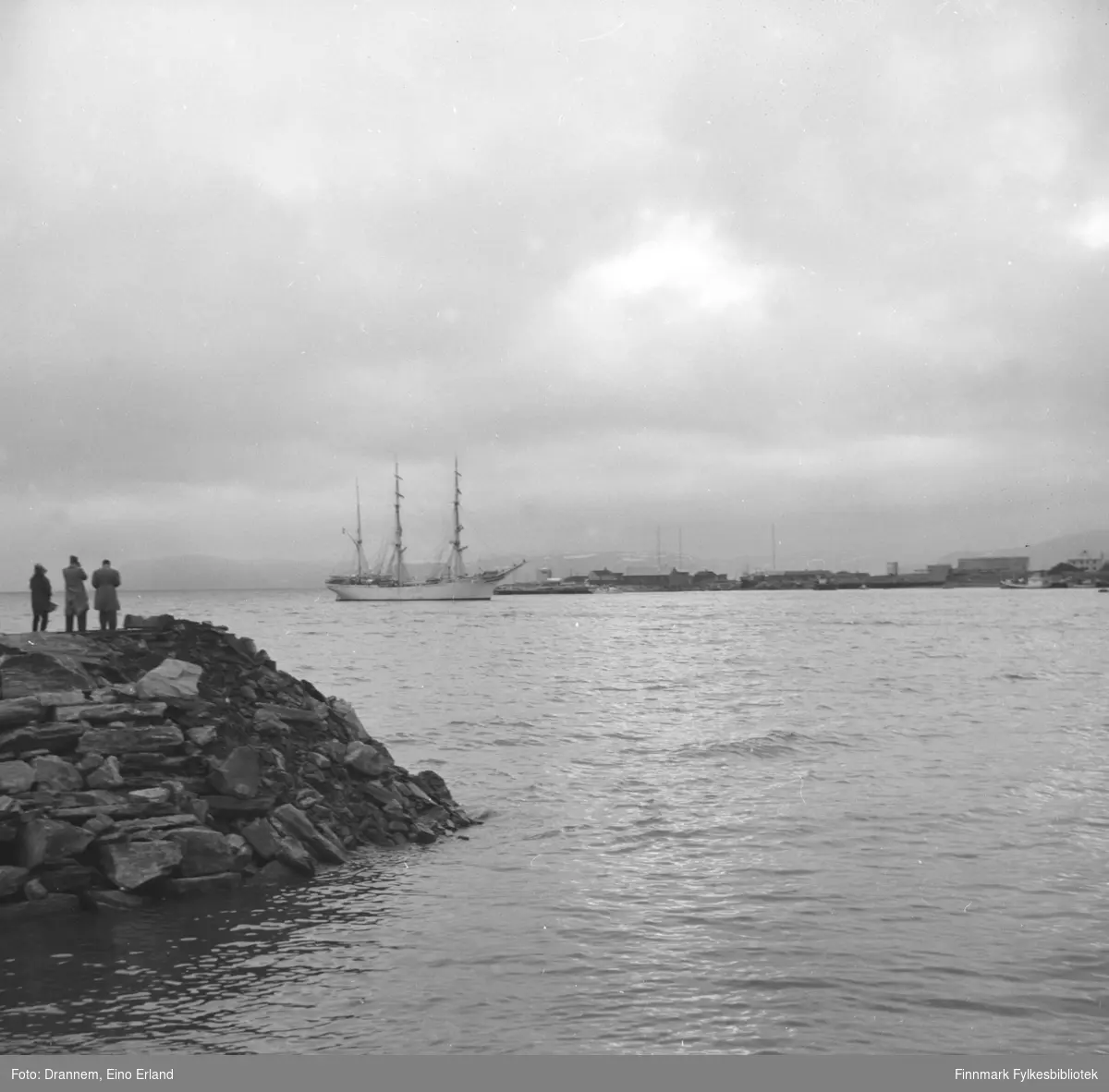 Utsikt mot Hammerfest havna. På venstre side av bildet ser man tre mennesker ståendes på moloen der de fotograferer og ser på skipet (med tre master og i hvit farge)