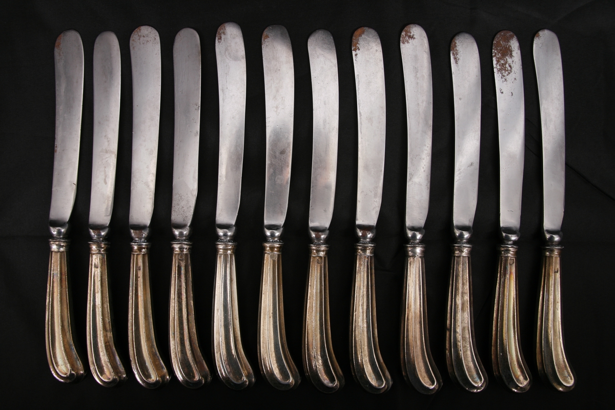 12 bordkniver med barokk-sølvskaft. Skaftet har et mønster av riller. Mot knivbladet går det en liten bue rundt skaftet. 
Over innskriften på selve bladet er det tre ulike symboler. Det ene ser ut som et kors, mens de andre er vanskelig å tolke.