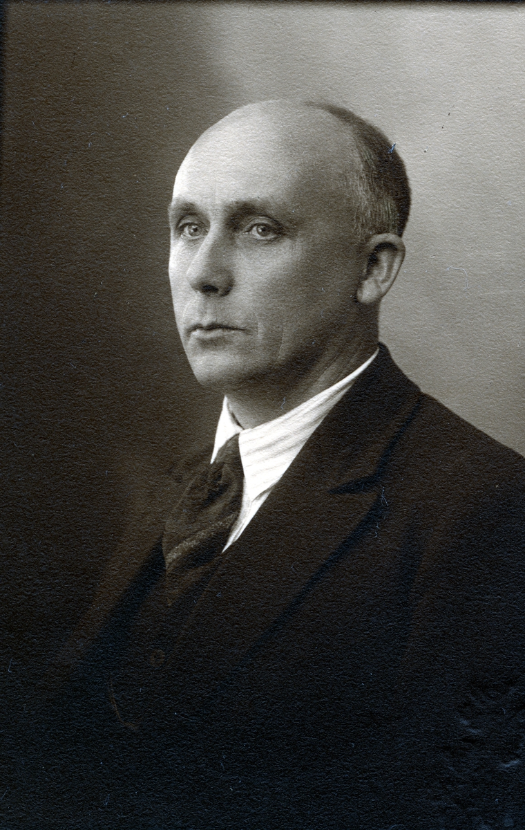 Portrett i halvfigur av mann kledd i mørk dress, slips og stripete skjorte.