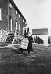 Eldre kvinne står ute på et tun, to bygninger i bakgrunnen.