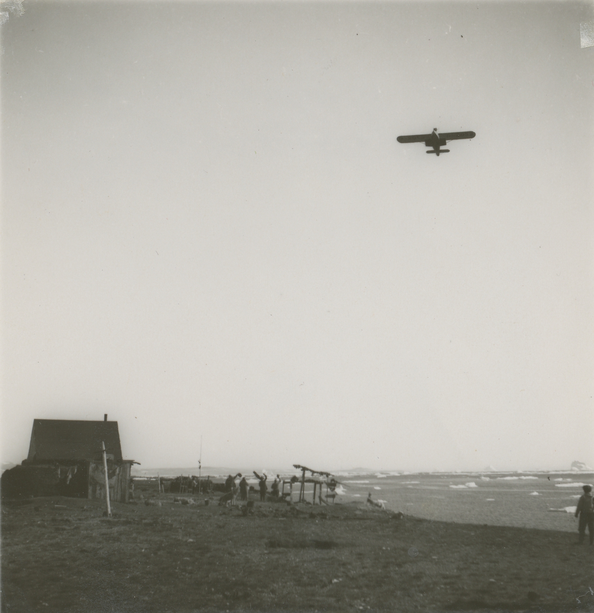 Wolfgang Von Gronau flyr over Kap Hope i Øst-Grønland. En gruppe mennesker står på bakken og vinker til flyet.