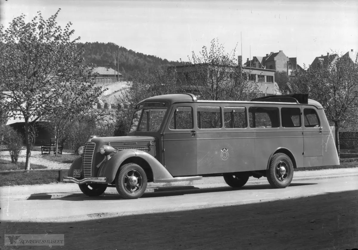 Buss fra Hjelset Auto med reg nr T-2755..T-2755 var en Diamond T, 1936-modell turistbuss med sammenfoldbart tak. Karosseriet ble påbygd av Ivar Røvik Karosserifabrikk, Røvik, og hadde bygg nr. 39. .Hjelset Auto ble startet i 1929 av Torstein Vorpenes, Hjelset, med ruter Hjelset-Molde. I 1943 ble Hjelset Auto overtatt av Haukebø & Rødseth, som også drev Aarø Automobilselskap. Hjelset Auto bygde ut lokalruter rundt Molde, Mordalruta fra 1946 og Ringbussen fra 1953. Trafikken på disse rutene vokste betraktelig utover 1950- og 1960-tallet. .I 1973 ble disse busselskapene slått sammen med Batnfjord Auto og Kleive Auto, og fikk navnet Molde Bilruter. Dette selskapet drev disse rutene til slutten av 1990-tallet. I 1972/1973 ble det bygd nytt anlegg for bussene i Fannestrandveien 71. Denne virksomheten har hatt tilhold der i alle år siden. I dag driver Vy Buss bussrutene, og Wist Last og Buss står for verksteddriften..I Romsdalsgata i bakgrunnen ser vi Ford-forhandleren Haukebø & Rødseths sin funkisbygning delvis skjult av løvverket.