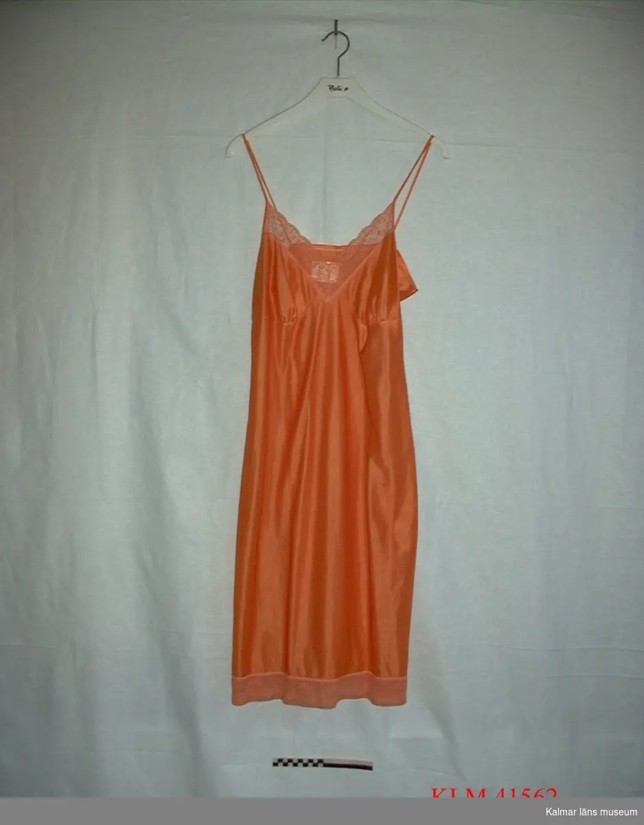 KLM 41562. Klänning, underklänning. Underklänning för dam av orange tyg, smala axelband, v-ringning fram, spets längs ringning och nederkant. Har använts under klänning KLM 41562.