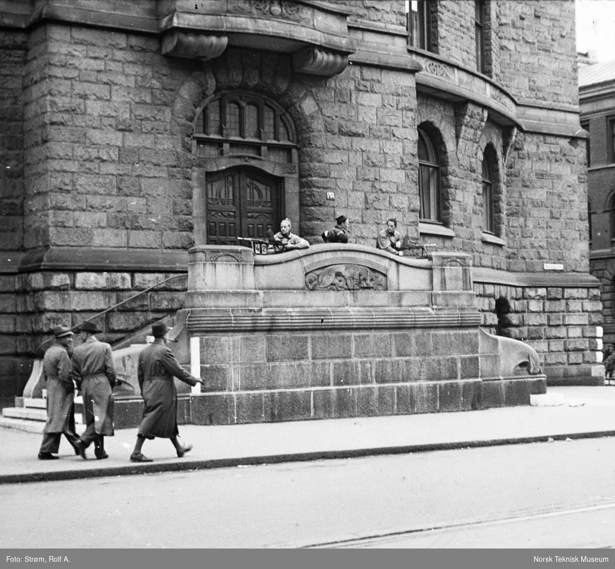 Finansdepartementet, tre bevæpnede milorgsoldater vokter inngangen, tre menn med hatt og frakk passerer, Oslo, mai 1945,