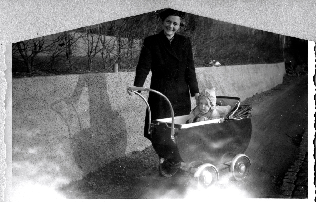 Rosa Pettersson (1912 - 1994, född Krantz) är ute på promenad med dottern Eva (född 1944, gift Kempe) som sitter i barnvagn, Stretered år 1945. 
Originalfotot är bränt och klippt efter en brand cirka 1945-1946 då makarna Pettersson hyrde en lägenhet i villa "Ljusbo", Kyrkbacken.