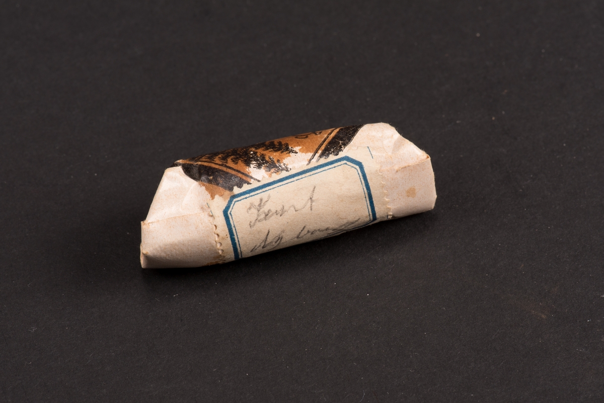 Rund begravningskaramell invikt i vitt papper. Dekorerad med en tryckt etikett som visar en gravsten med urna flankerad av fyra granar. 
På baksidan en etikett med ett namn "Kurt Nyborg".