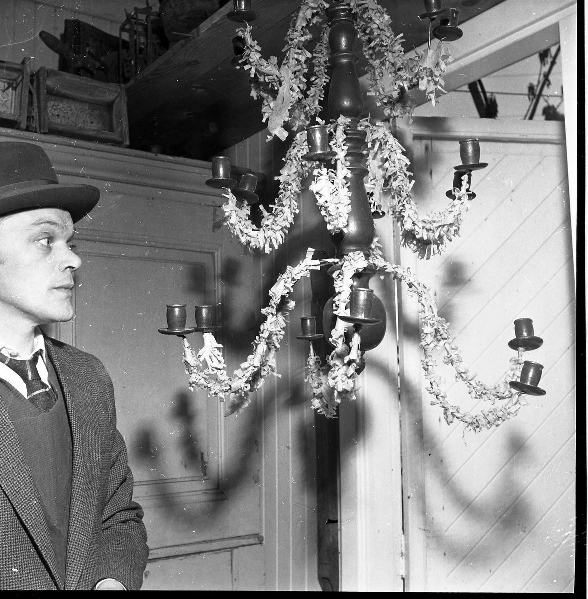 En man med hatt står vid en upphängd med fransat papper dekorerad ljuskrona.
