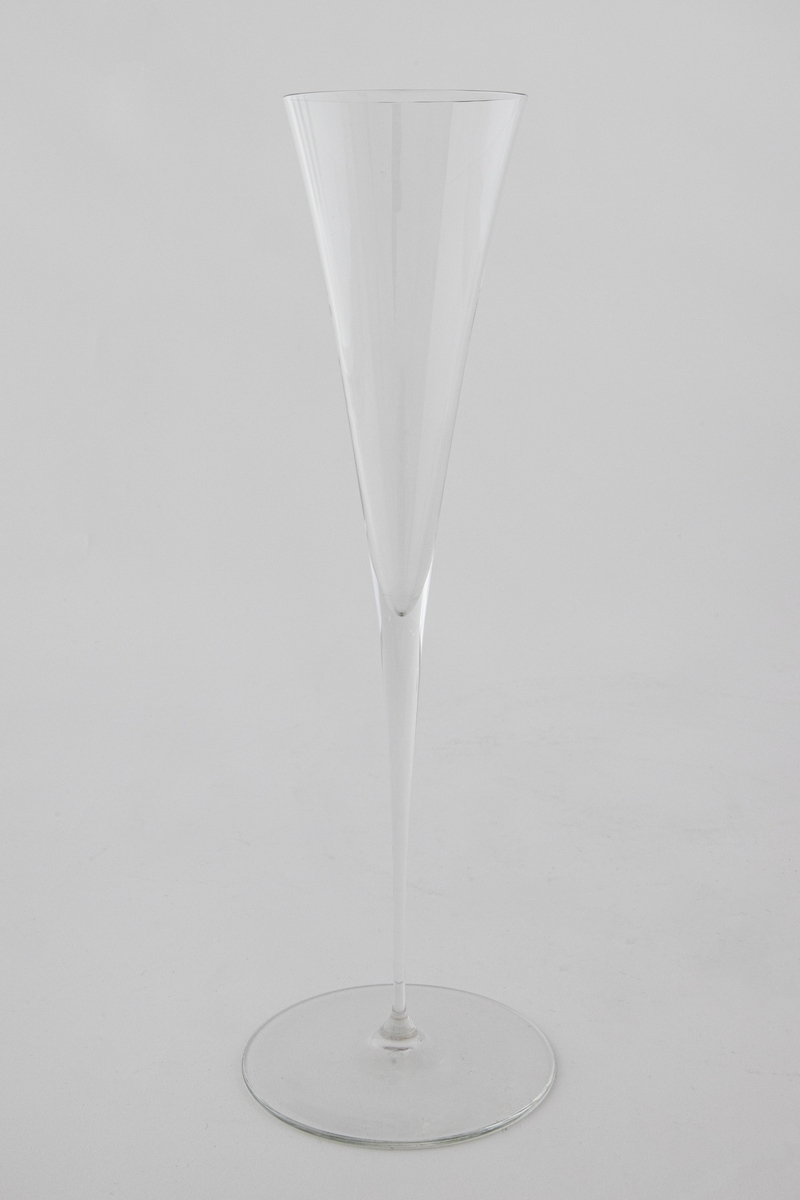 Høyreist champangeglass i klart glass. Konisk kupa som glir over i en stett, som hviler på en sirkulær fotplate.
