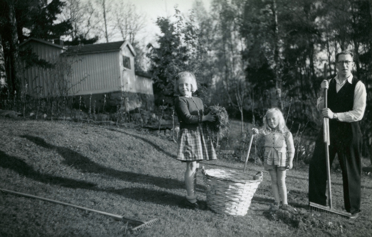 Systrarna Eva (född 1944) och Karin Pettersson (född 1947) hjälper pappa Bror "Petter" Pettersson (1913 - 1984) att samla löv vid familjens bostad på Gamlehagsvägen 17 i Torrekulla, cirka 1951. I bakgrunden till vänster ses Danielssons sommarstuga.