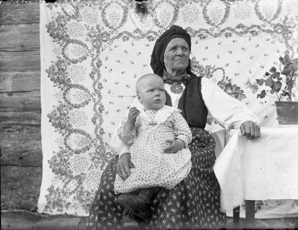 Portrett av eldre kvinne i folkedrakt fra Vest Telemark, med barn på fanget.

Fotosamlingen etter Olav Tarjeison Midtgarden Metveit, (1889-1974), Fyresdal. Senere (1936) kalte han seg Olav Geitestad.