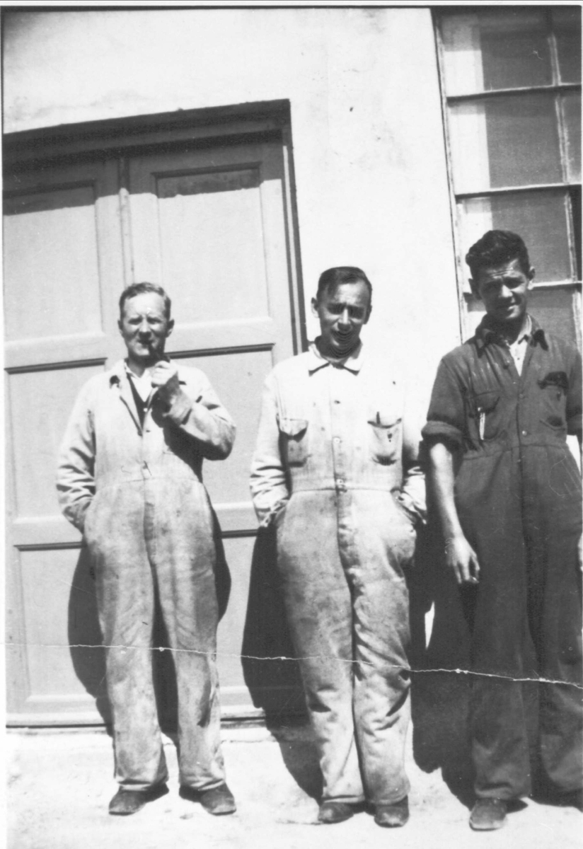 Tre menn i arbeidstøy stående utenfor et verksted.
