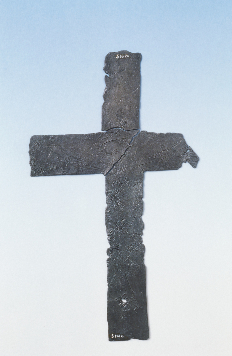 "1616. Et blykors, hvis ene side er forsynt med en korsfæstet Kristusfigur og den anden med runer."   Dateringen bygger på stilistiske kriterier i Kristus-fremstillingen.  Korset har ganske omfattende overflatekorrosjon.