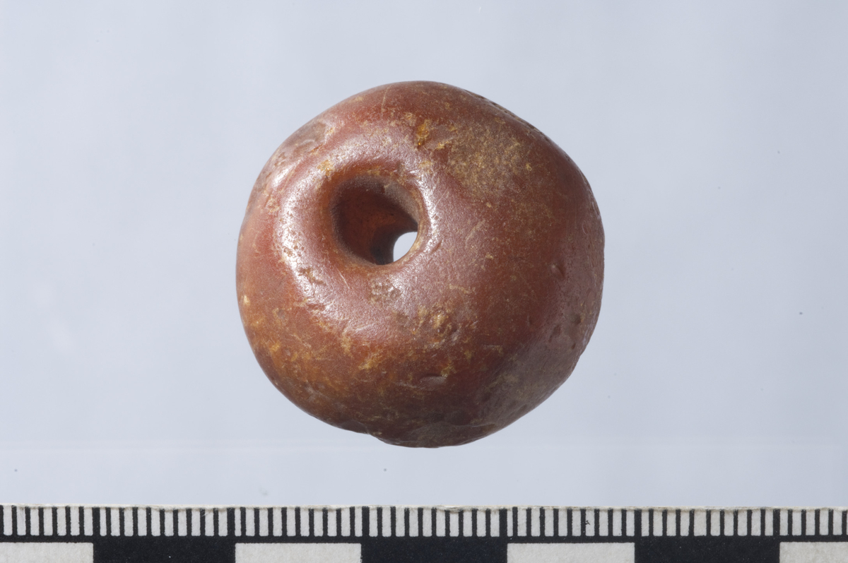 "Større perle av rav, rimeligvis fra vikingetiden, sl. Jan Petersen: Vikingetidens smykker. Fargeplansje fig. 5. 2,7 cm. i tverrmål, 1,8 cm. tykk." Ny beskrivelse etter revisjon: Perle av rødbrun, flammet rav, avrundet med en konkav side og en sterkt hvelvet og skjev side. L: 11-18 mm, diam: 27-28 mm. Perlen er noe skadet i overflaten.