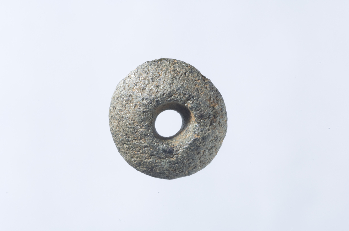 d) En perle av kleber(?). Perla er rund, sideflatene rygget, hullet er boret fra begge sider og har derved en bikonisk form. Diam. 14 mm, t. 7 mm, hullets diam. 6 mm.