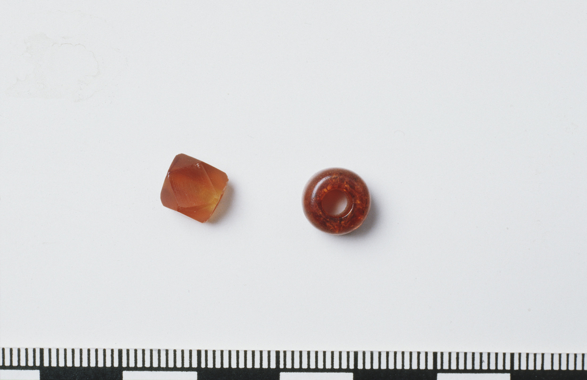 Mørk, oransjerød perle av rav, gjennomskinnelig, marmorert, noe krakelert. Avrundet, med plane, parallelle sider, stL/D: 6,5/8,5 mm. D: hull 3 mm. Fnr. (F.): 387.