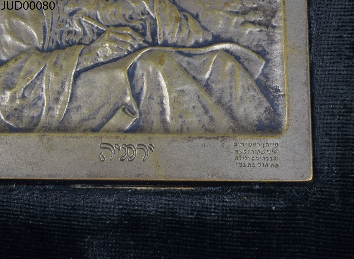 Svart sammetstavla med rektangulär mässingsplakett i mitten, föreställande skrivande man. I mässingsplattans nedre högra hörn är hebreisk text skriven.