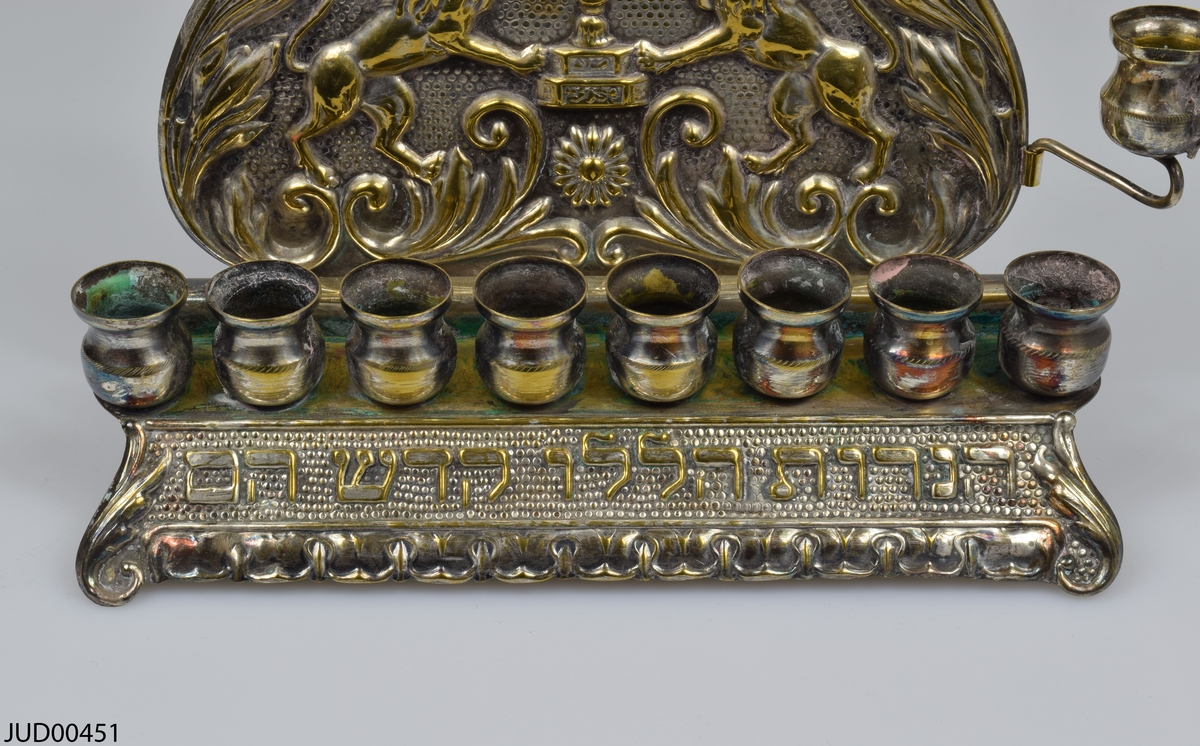 Chanukaljusstake tillverkad av silveröverdragen mässing. Dekorerad med en krona, hebreisk text samt en menora flankerad av två lejon. Bakstycket är fastsatt mot botten med skruvar i bakkant.