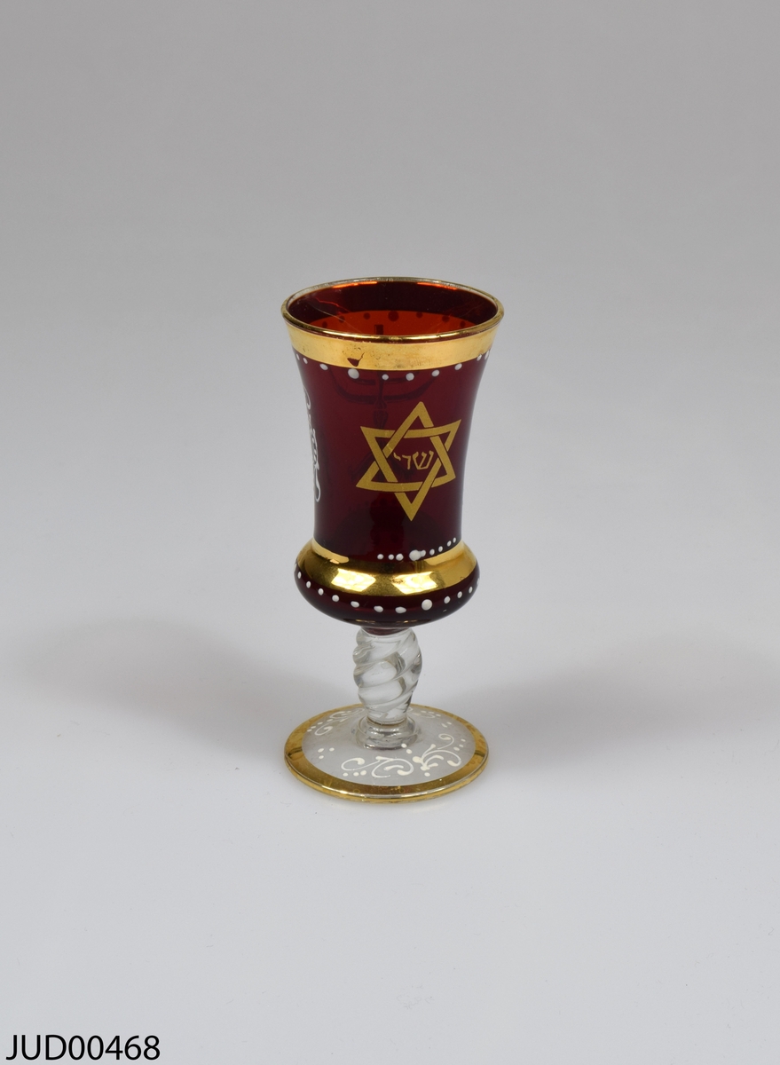Glasbägare, med genomskinlig fot och rödfärgad cupa. Dekorerad med en guldfärgad menora och en davidsstjärna med hebreisk text i. Även vit snirklig dekor.
