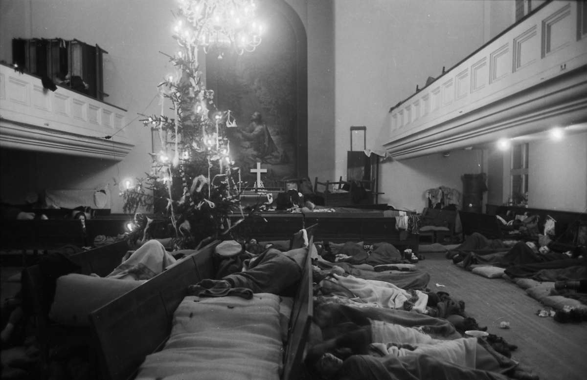 Tvangevakuerte fra Porsanger sover i Metodistkirken i Trondheim i jula 1944. De ligger på madrasser som er tett plassert på kirkebenker som er plassert sitteflate ved sitteflate, men også tett i tett på gulvet. De fleste, men ikke alle, har enkle pledd å dekke seg til med. Noen personer følger med og vender ansiktene sine mot fotografen. Man ser kirkerommets gallerier og møbler som er stablet opp, både nede og oppe. I bakgrunnens midte ser man prekestolen, alteret og altertavla. Likevel er det juletreet som dominerer bildet. Det er pyntet med lys og norskeflagg. Over treet henger det en lysekrone. På gulvet kan man ser en nattepotte og papir, samt til tørking utstoppede skaller.