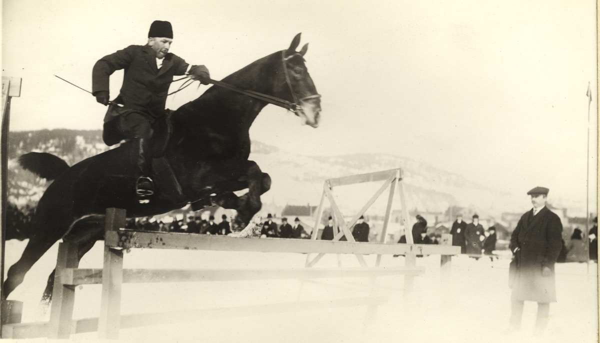 Westye P.  Egeberg på hesten "Njål" forserer et hinder under et ridestevne på Marienlyst i Drammen. De vant førstepremie. Tilskuere står i bakgrunnen. Fotografert 1917.