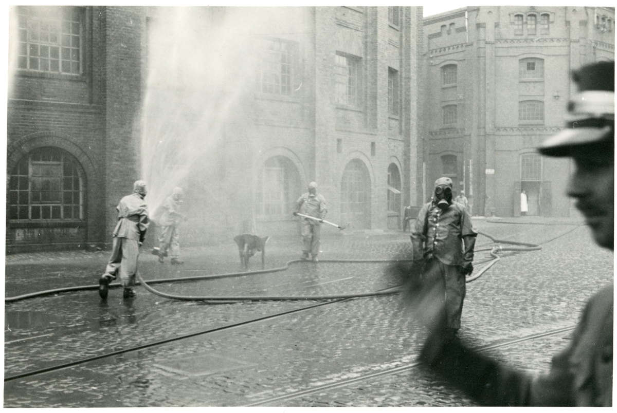 Brannøvelse i fabrikk i Hannover, Tyskland. Menn med gassmasker styrer brannslangen. En mann i uniform styrer øvelsen i forgrunnen. Fotografert 1935.