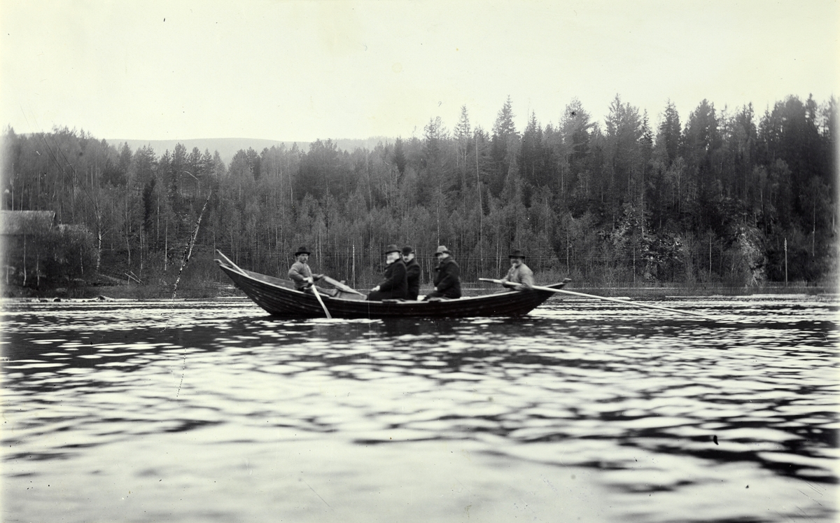 Tre menn, antagelig på befaring fra Christiania Tømmerdireksjon, i robåt med to roere på vei nedover Glomma ved Barkald i Alvdal. Fotografert i 1907.