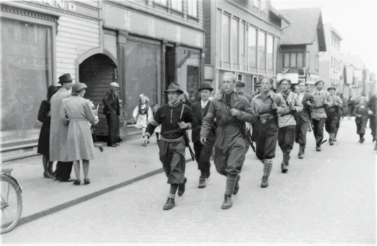 Feiring av freden 1945. Hjemmestyrkene marsjerer nord i Strandgaten 7. eller 8. mai. Tilskuere på fortauet og i gaten. Husrekke til venstre.