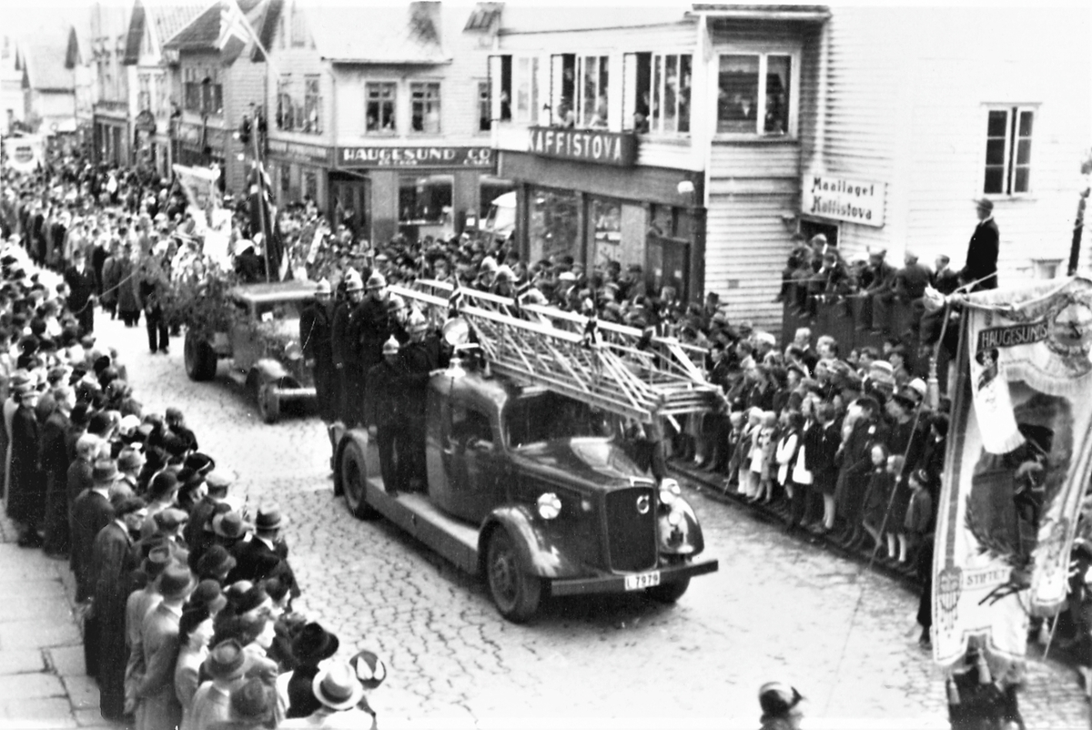 Feiring av freden 1945. Som et nytt innslag i folketoget 17. mai, deltar her byens brannvesen med sine biler og sitt mannskap. (Husrekke med "Kaffistova" til høyre).