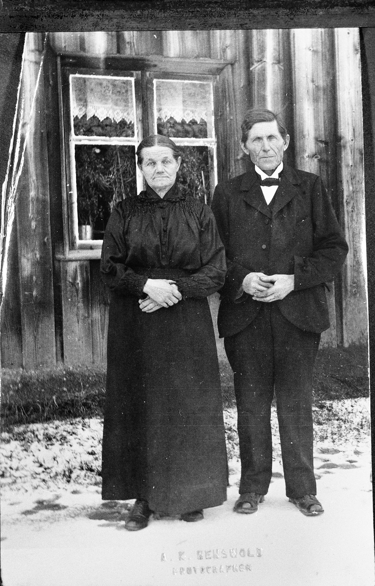 Karine og Emil Hagen står stivpyntet foran kjøkkenvinduet i Rognstadhagen. Gardinene set ut til å være sydd av hvite kongresgardiner, og påsydd hjemmeheklede bord-spisser.
