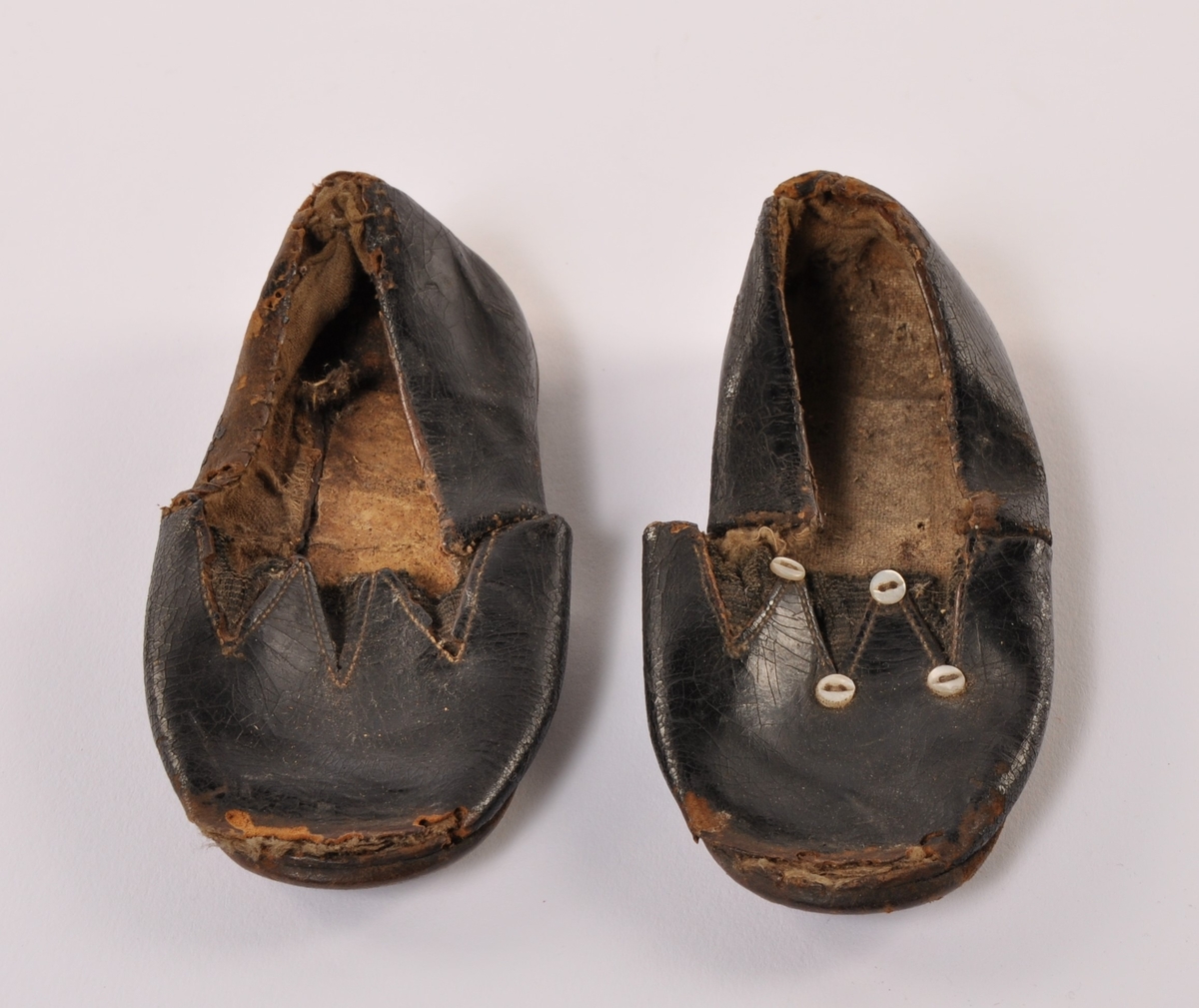 Et par barnesko i sort skinn med lærsåle, med strikk over vrist. Ene skoen har 4 stk pynteknapper i perlemor, mangler på den andre.