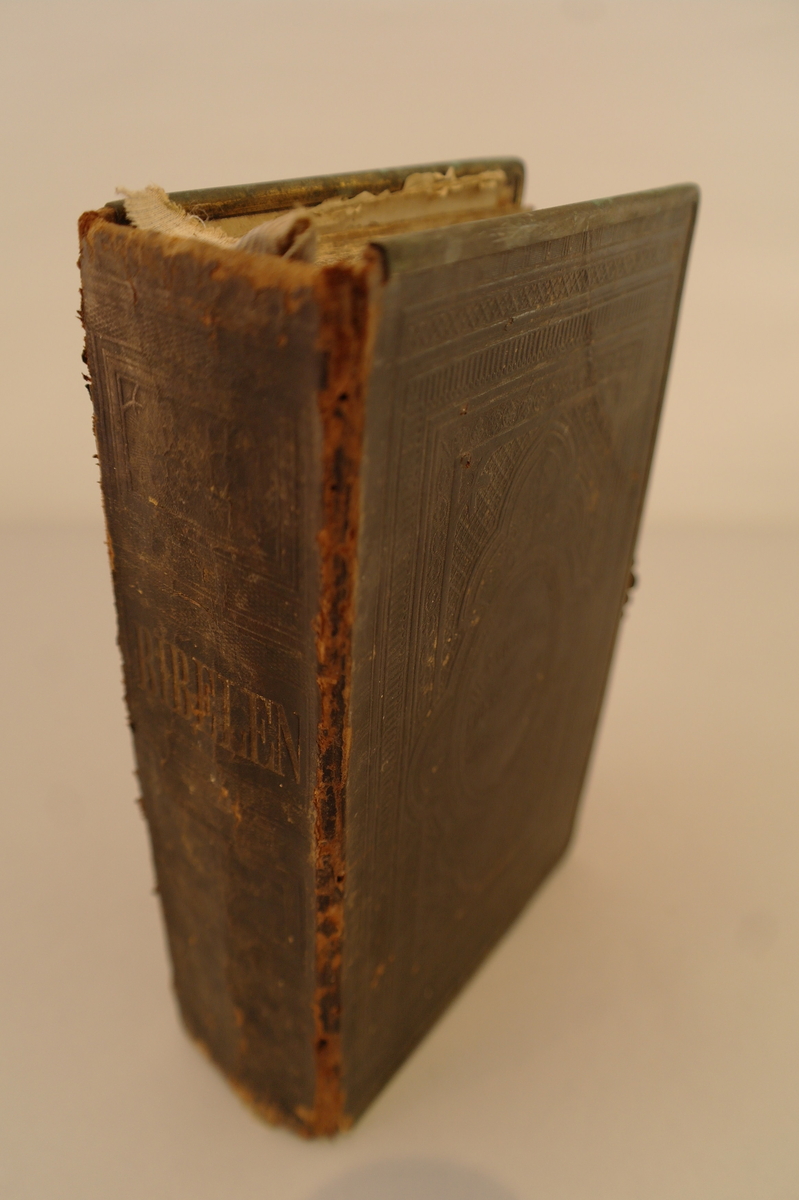 Bok: Bibelen. Det Britiske og Udenlandske Bibelselskab. Svart bokomslag med metallbeslag rundt kantane og en metallspenne for å lukke boken.