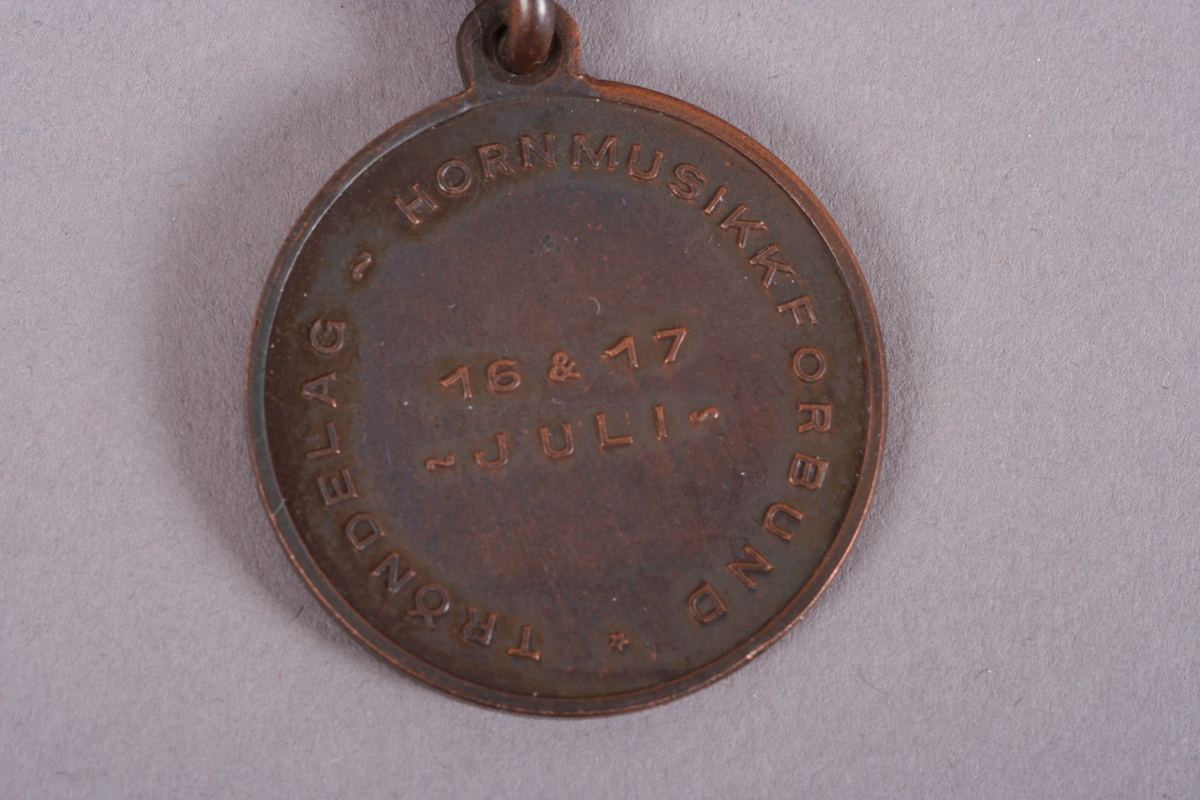 Medalje med påskrift Røros janitsjarorkester, 1949.
På forsiden motiv fra Røros sentrum med klokke og kirketårn.

På baksiden påskrevet: Trøndelag Hornmuiskkforbund 16 & 17 juli.