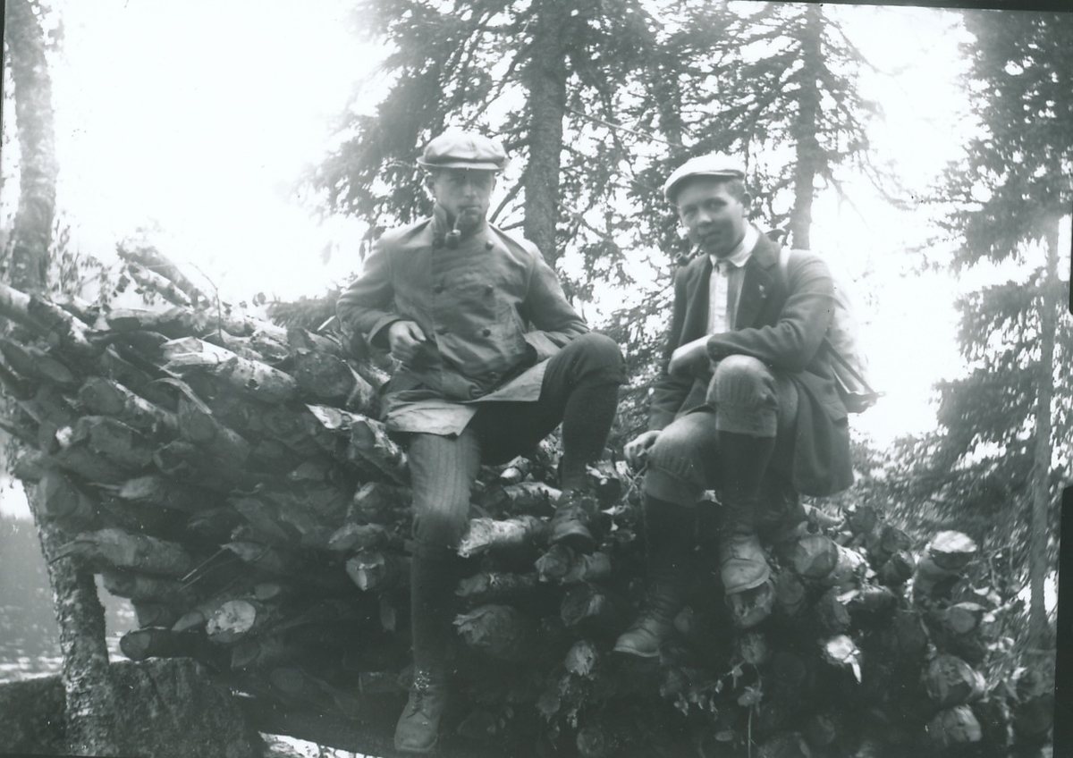 Harald Rydgren til høyre sammen med en ukjent ung mann. De sitter på toppen av stabel med trestokker eller staur i et skogslandskap. Begge har skyggeluer, sportsantrekk og piper.