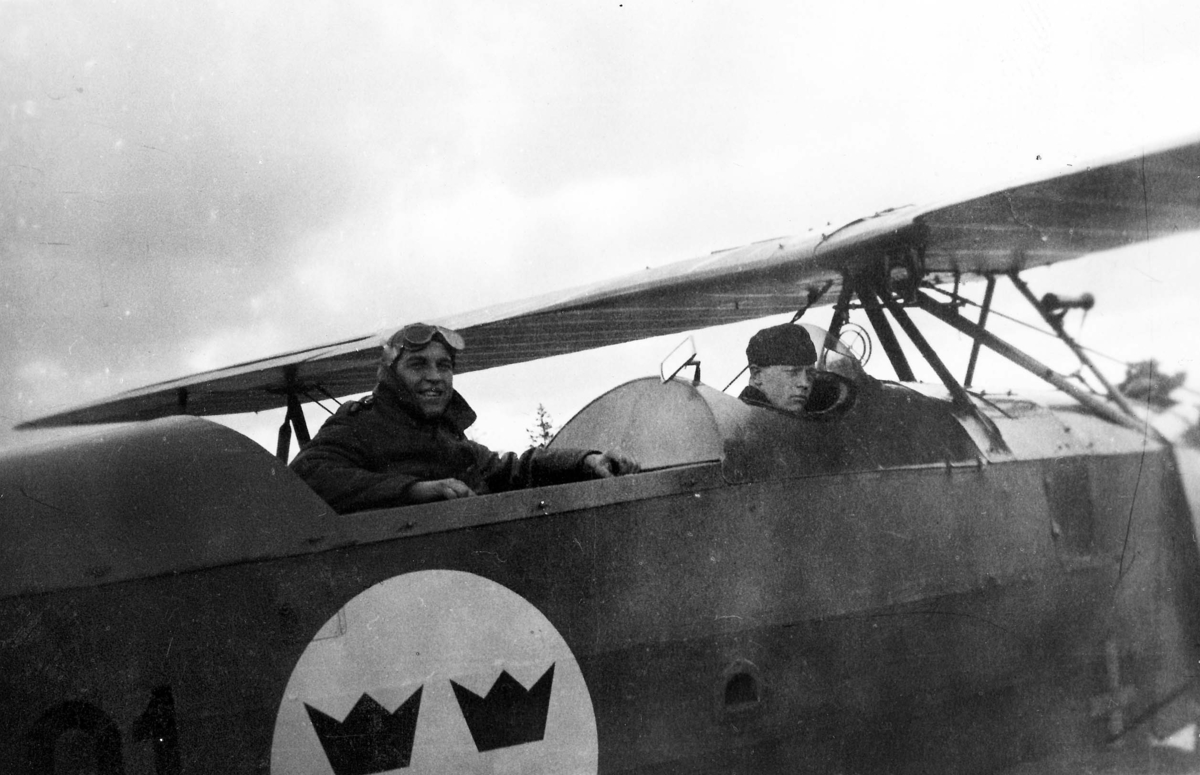 Två militärer sitter i flygplan T 1. En är klädd i flygdräkt. Vy bakifrån. 1930-tal.