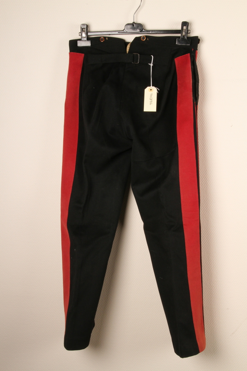 Uniformsbukse i svart ullstoff, med to brede, røde renner i hver side. Spensel i midjen midt bak. 