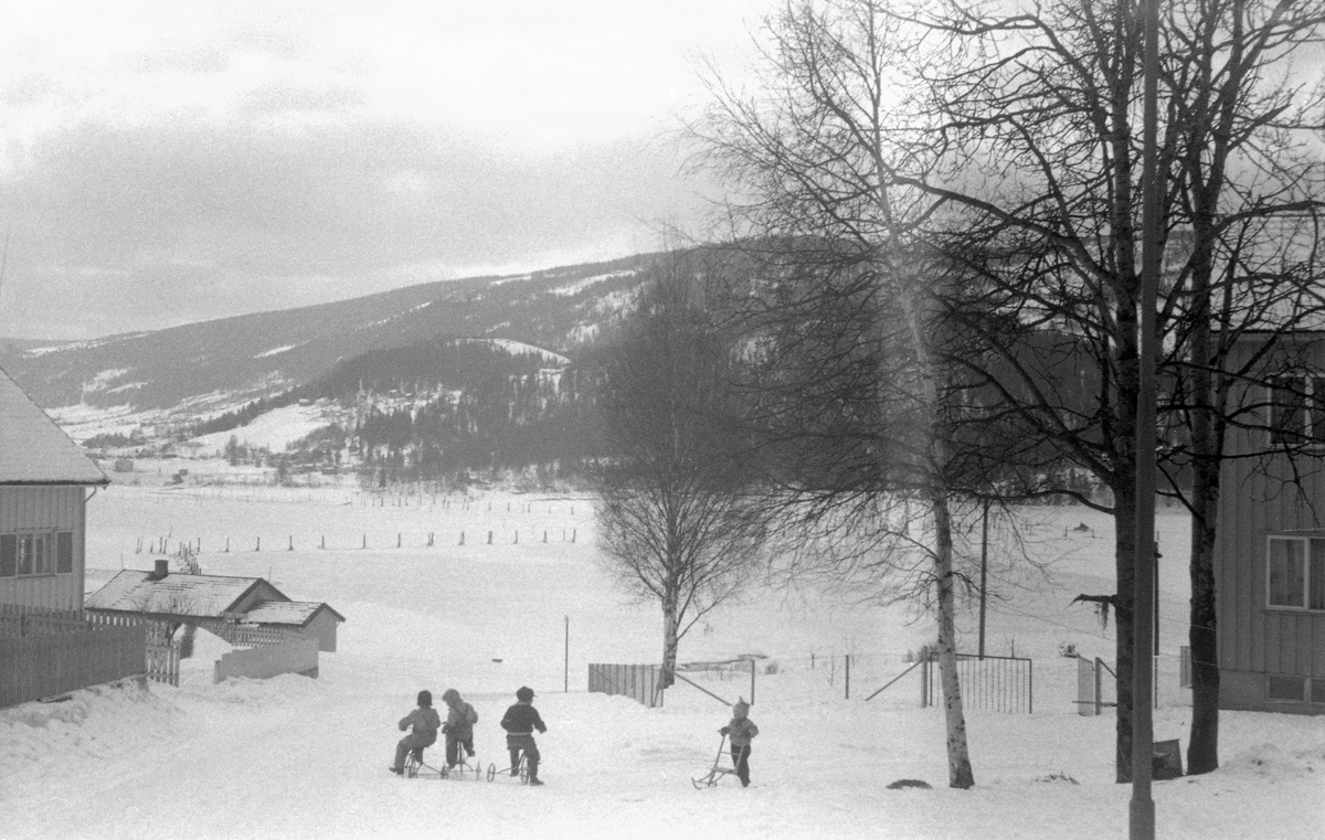 Vinterbilde fra Lillehammer i midten av 1950-åra. Bildet er tatt fra en veg som førte ned mot Gudbrandsdalslågens utløp i Mjøsa. I forgrunnen ser vi fire barn som lekte i vegen, tre med trehjulssykler og ett med spark, Ved bildets ytterkanter (både til venstre og høyre), ser vi bolighus som var bygd langs den nevnte vegen. I mellomgunnen lå det is- og snødekte vassdraget, der pælene i lenseanlegget stakk opp. På motsatt side av elva ser vi Pinnikhaugen med Vottestadhøgda i bakgrunnen.