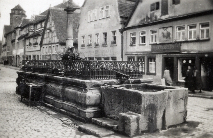 En gammal stenbrunn mitt i en gata. Till vänster syns en pelare (med trol. en helgonbild) och i vänstra hörnet ett torn. 
Text under fotot: "Motiv från Tyskland, 1931". (Nürnberg?).