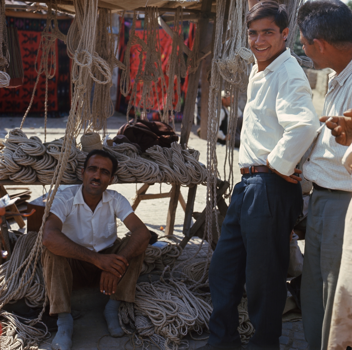 På marken framför ett försäljningsstånd med rep sitter en man på marken. Bredvid honom står två män. En av dem ler in i kameran.