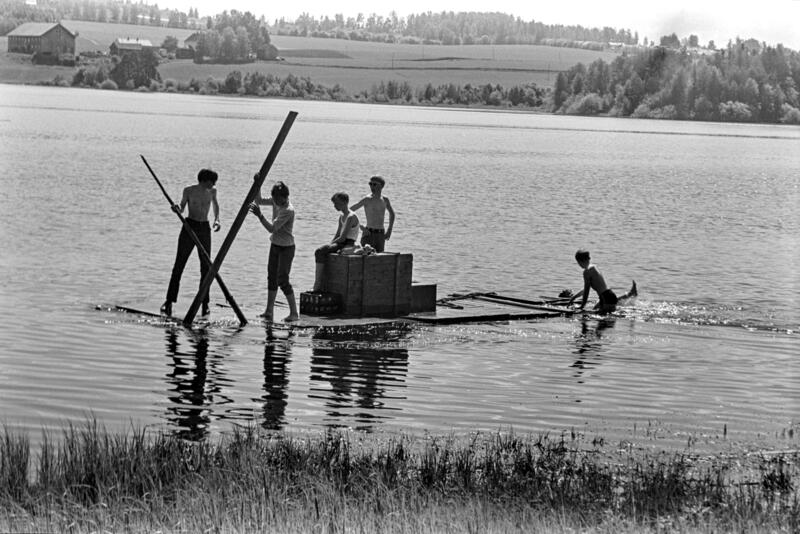 Svart-hvitt bilde av fem ungdommer i shorts som seiler på en hjemmelaget flåte. (Foto/Photo)