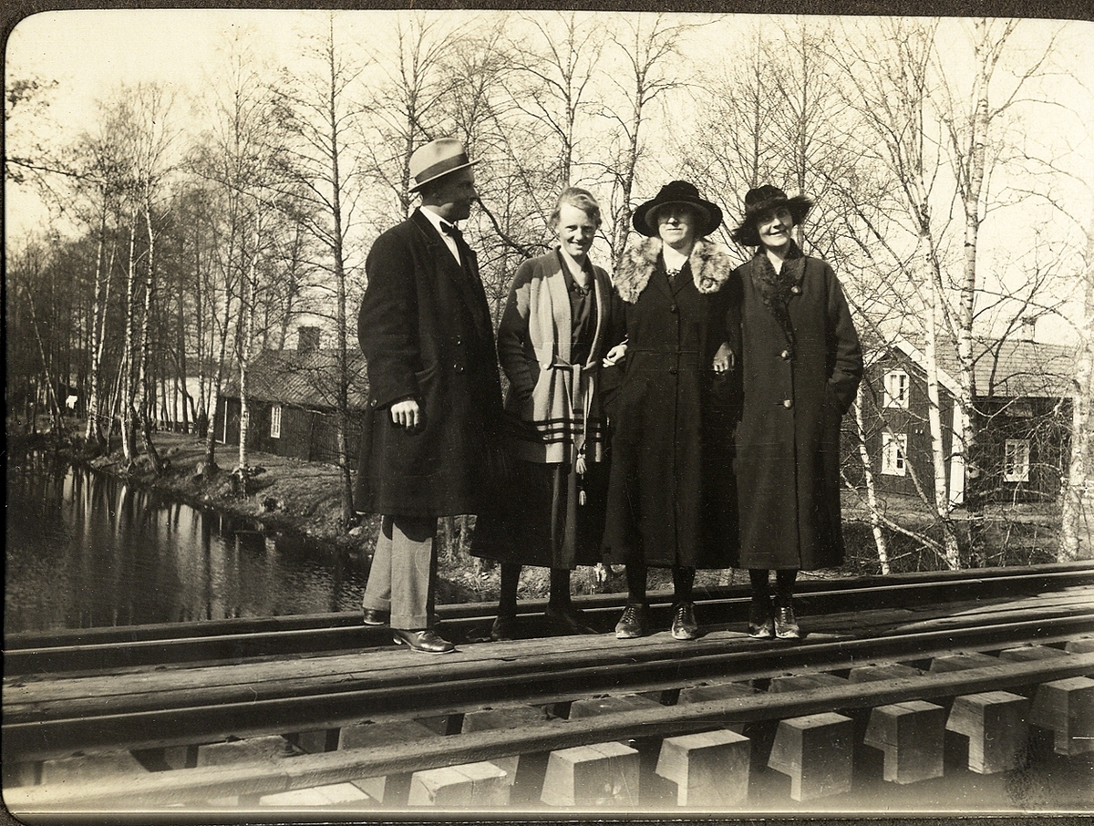 Tre kvinnor och en man står på en järnvägsbro (?) över en å.