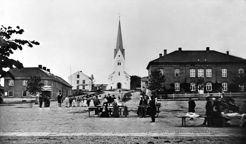 Svart-hvitt fot som viser et torg med handelsboder og torgkoner i "gammeldagse" klær, et hus i hver bildekant og Hamar domkirke bakerst i midten. (Foto/Photo)