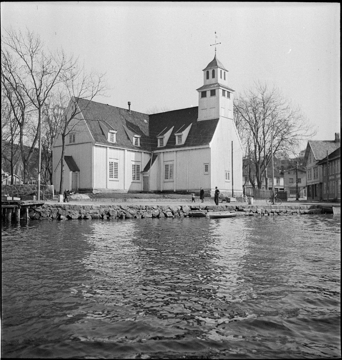 Egersund kirke sett fra Vågen og fra byen. En gruppe menn sitter ved inngangspartiet.