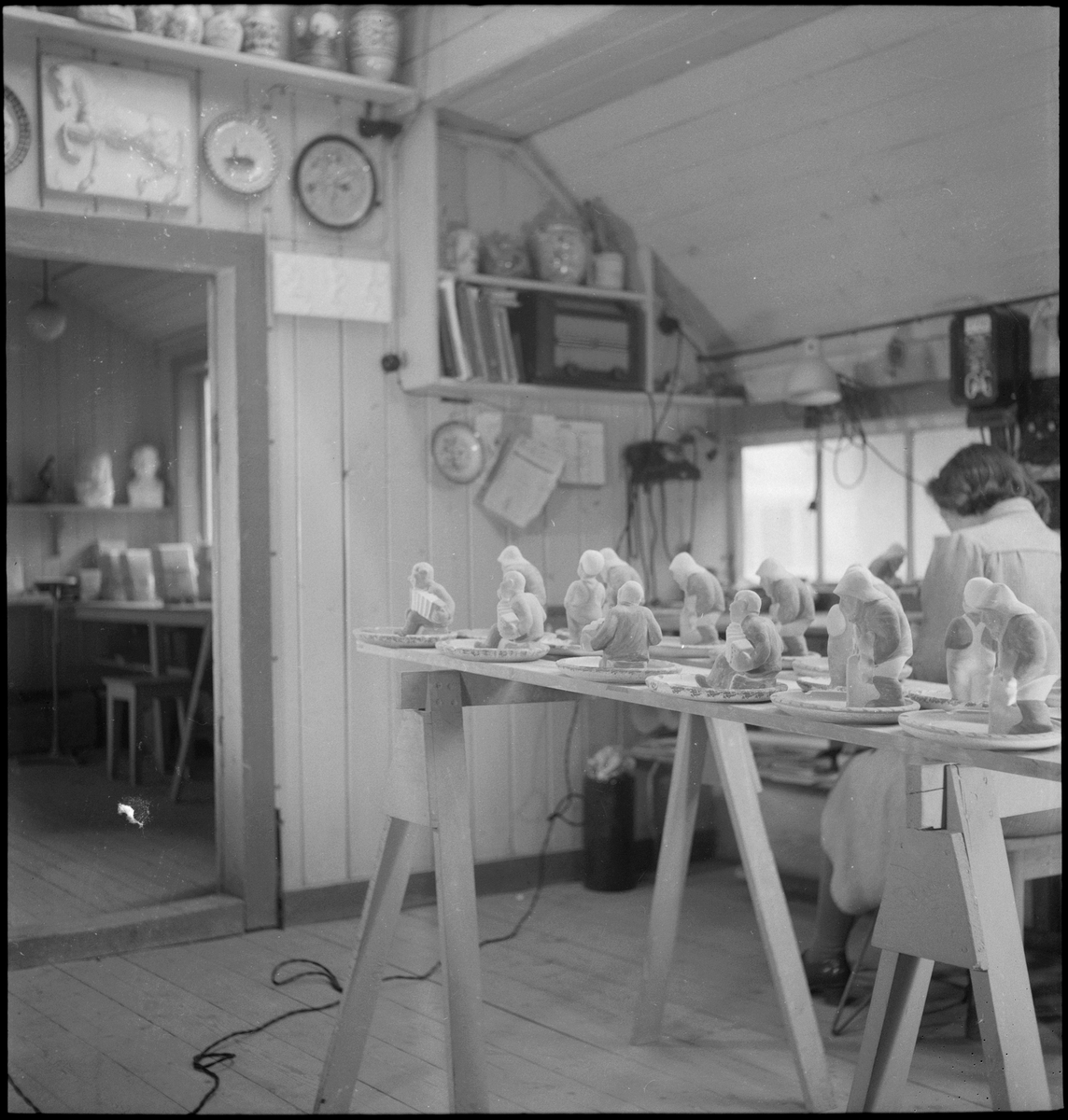Keramiker Thorvald Olsen og en kvinne maler keramiske produkter i Olsens verksted. På bordet er det flere keramikkfigurer.