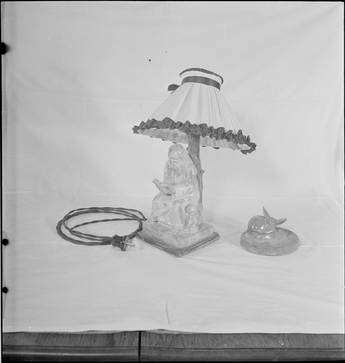 Keramiske produkter laget av keramiker Osvald Osmundsen. Det er en lampe formet som en elefant og en lampe formet som en mann som leser under et tre og noen keramikkfigurer.