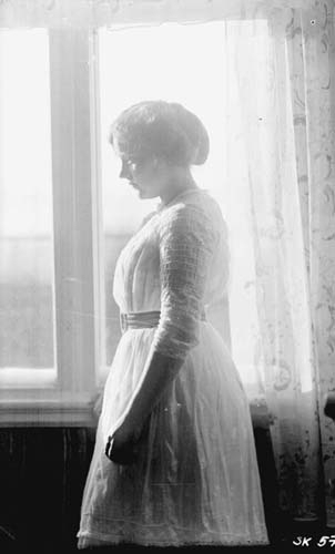 En kvinna i ljus klänning står i profil inomhus vid ett fönster. Solen skiner in från vänster.